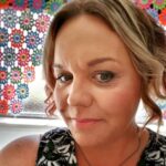 Close up of smiling woman with blonde hair in front of colourful crocheted flower curtain